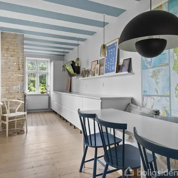 Langt, lyst rum med hvidt og blåt loft. En reol med billeder og planter langs væggen. Kort over Danmark dekorerer væggen. Blå stole ved spisebord. Vindue bagtil.
