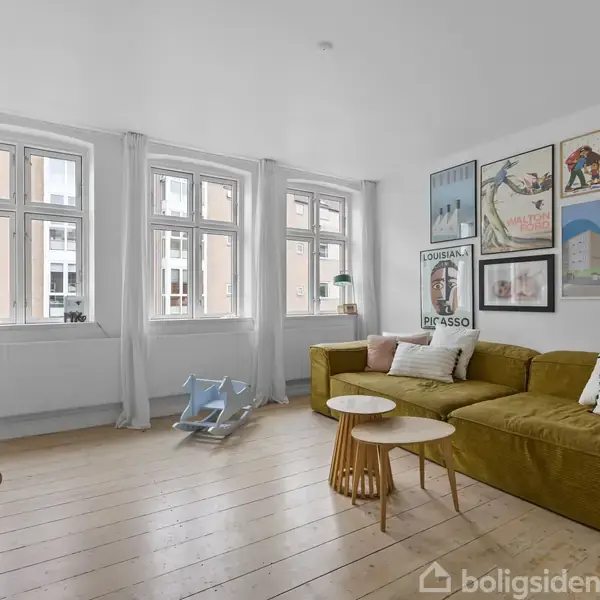 En stue med en gul sofa under flere vægbilleder, en lænestol til venstre, og et lille hvidt bord. Vinduer med hvide gardiner kigger ud mod bygninger.