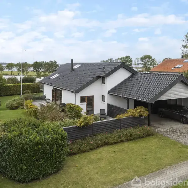 Et hvidt hus med sort tag står i en velplejet have med buske og træer. Der er en carport med en parkeret bil. Naboer ses i baggrunden.