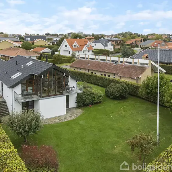 Et moderne hus med store vinduer og sort tag ligger i en grøn, velplejet have. Huset har en hævet terrasse. Omkringliggende område indeholder flere huse i en boligudstykning.