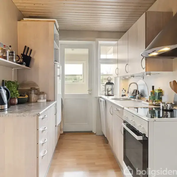 Køkken med træskabe og -gulv. En bordplade med elkedel og grøntsager, ovn og køkkenredskaber. Dør fører til lys rum. Spisebord i baggrund.