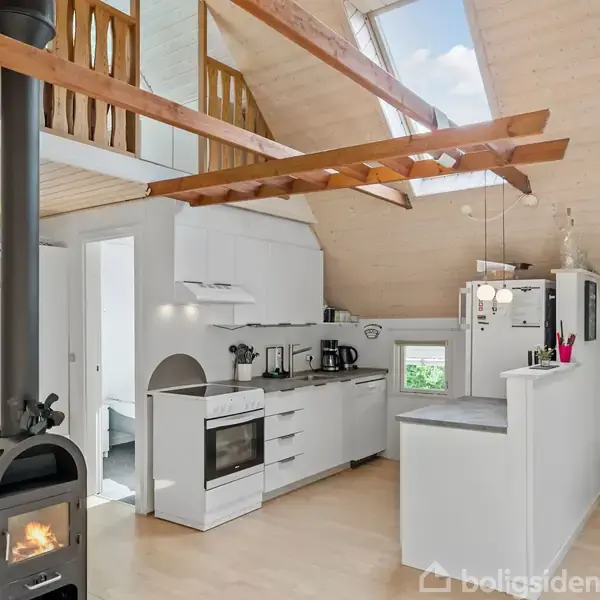 Brændeovn står tændt, ved siden af et moderne hvidt køkken med indbygget ovn og emhætte. Træbjælker og skråt loft med ovenlys; rummelige, lyse omgivelser.