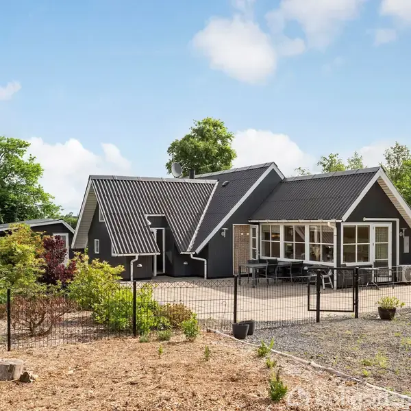 En moderne, sortmalet hus med asymmetriske taglinjer står omgivet af grønne træer og buske. Et hegn omgiver gårdspladsen, hvor der er et grusbelagt område foran huset.