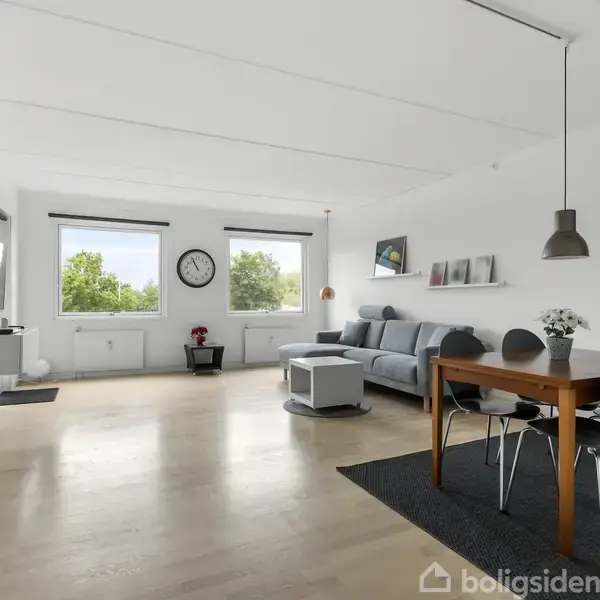 En lys stue med en grå sofa ved væggen. To vinduer giver naturligt lys. Et spisebord med grå stole står til højre. Vægur hænger mellem vinduerne.