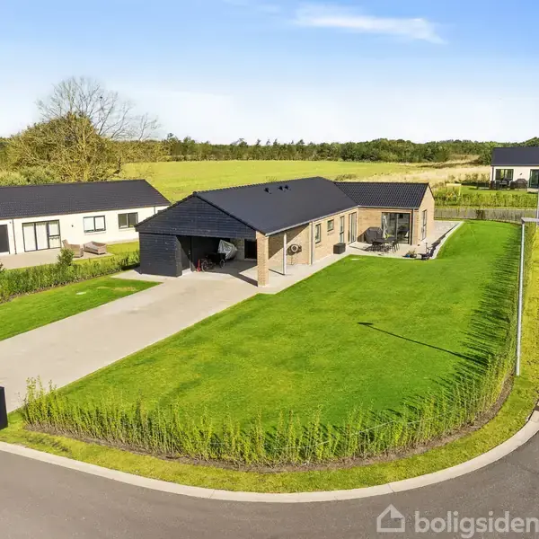 Et moderne, en-etagers hus står på en velplejet, grøn græsplæne med en flisebelagt indkørsel og carport. Omkranset af andre huse i et fredeligt boligområde.