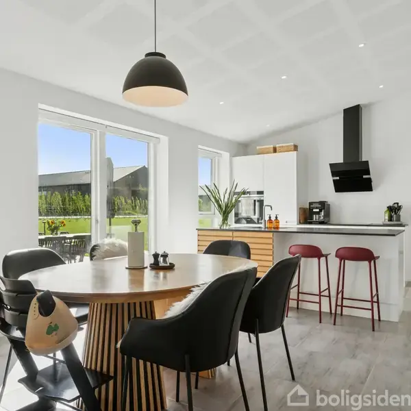 Rundt spisebord med stole i en moderne spisestue. Store vinduer giver udsigt til en grøn have. Væggene er hvide, og der er et køkkenområde med barstole og planter.