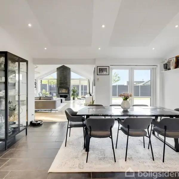 Spisestue med et langt bord, seks stole og en vase med blomster på. Glasvitrine til venstre, stort ur på væggen. Rummelig stue i baggrunden med kamin og store vinduespartier.