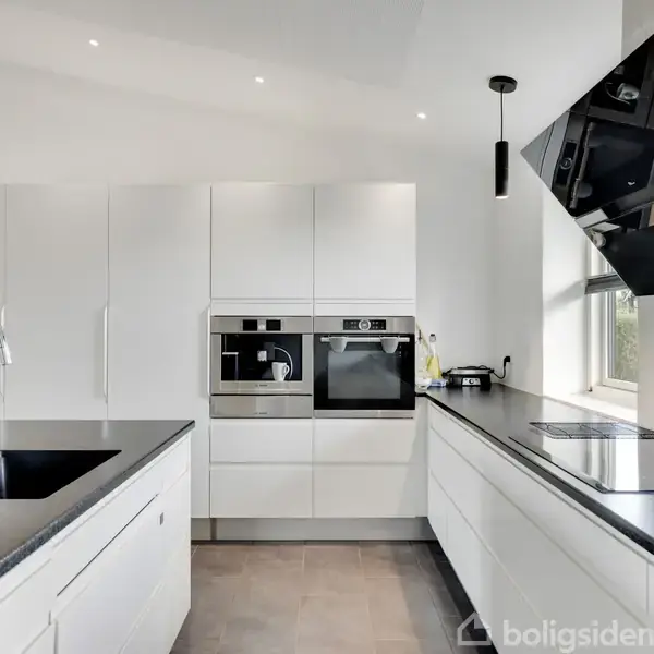 Et moderne køkken med hvide skabe og indbyggede ovne. En sort bordplade indeholder en vask og komfur. Rummet er lyst op af naturligt lys fra et vindue ved siden af emhætten.