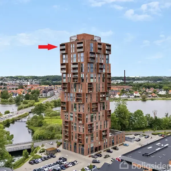 En højbygning i rød mursten står stille ved en flod, omgivet af træer og bygninger. En rød pil peger på øverste etage.