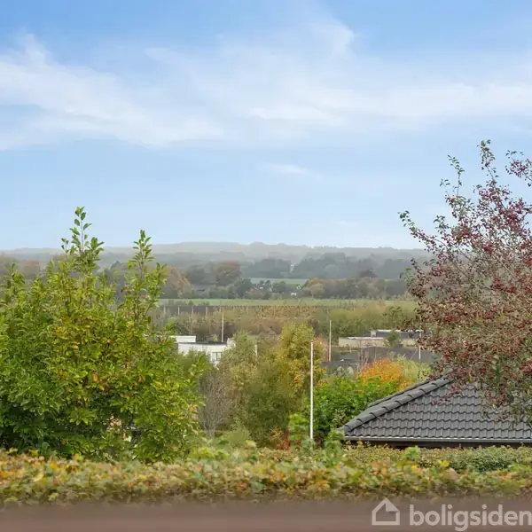 En have med grønne og rødlige træer omgiver et tag i grå tegl. I baggrunden ses en åben udsigt over marker og skov under en blå himmel.