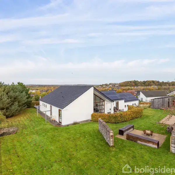 En hvid bygning med sort tag står stille i en grøn have. I haven er der træer, buske og en betonstruktur. Det er en klar dag med få skyer på himlen.