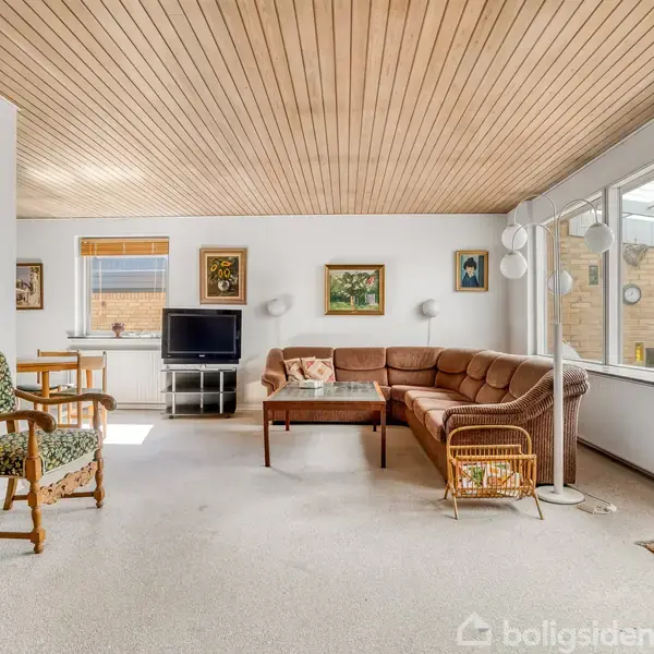Brun lædersofa står i en stue med tæppebelagt gulv, omgivet af træmøbler og malerier på væggene. Sollys strømmer ind gennem store vinduer, og der er en lampe i hjørnet.