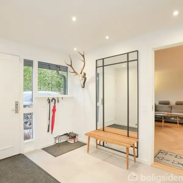 Entré med tøjstativ, hvor en jakke hænger, og specielle lampeformede lysestager på gulvet. Paraply står ved siden af døren. Lyse vægge og et vindue med udsigt til træer.