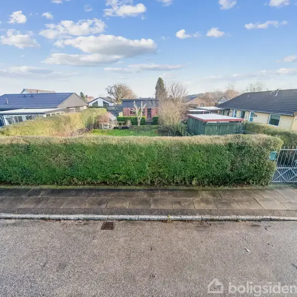 Hæk omkranser en have, som ses fra vejens perspektiv. Rækkehuse i baggrunden under en blå, let skyet himmel skaber en stille, beboelig atmosfære.