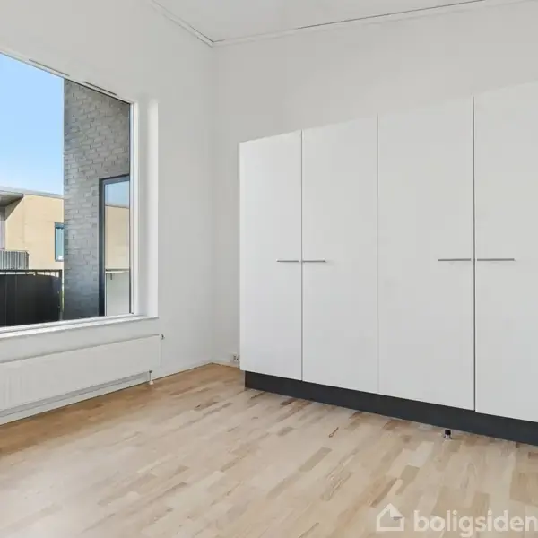 Skab står stille i et minimalistisk, lyst værelse med lyse trægulve. Store vinduer viser en udsigt til et moderne boligområde. Radiator placeret under vinduet.