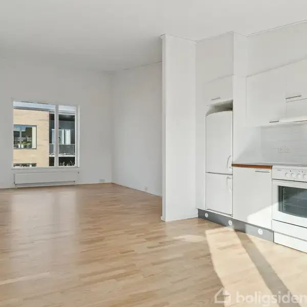 Et køkken med hvide skabe og komfur står i et lyst rum med trægulv. I baggrunden ses et stort vindue og en radiator.