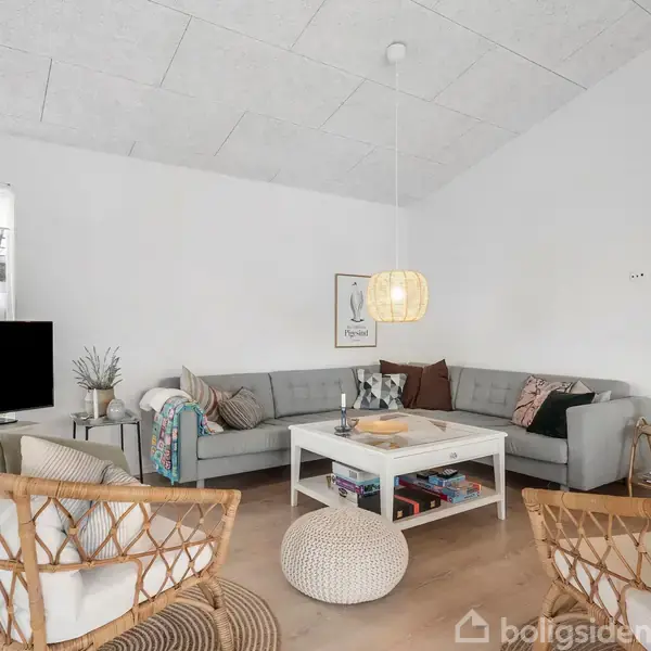 Grå hjørnesofa med puder omkranser et hvidt sofabord med bøger i en stue. To kurvestole og en lampe hænger over bordet. Fjernsyn og planter ved vinduet.