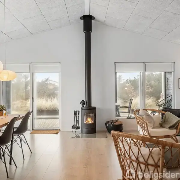 Brændeovn står i midten, brændende, omgivet af lyse træmøbler i en moderne stue med store vinduer og hængende lamper, omgivet af natur udenfor.
