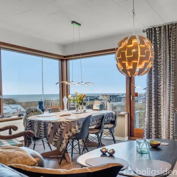 Stue med spisebord dækket til seks personer, placeret foran store vinduer med havudsigt. Over bordet hænger en moderne lampe og rummet er indrettet med sofaer og dekorationer.
