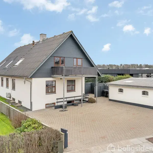 Et to-etagers hus med skifertag og en lille balkon ses. Huset er placeret ved siden af en garage. Baggrunden har en grøn græsplæne og buske. Himlen er let skyet.