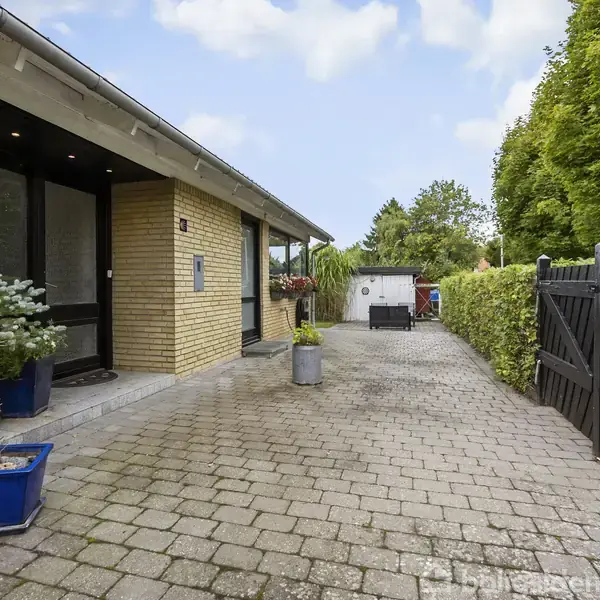 En gul murstensbygning med en indgang prydet af blå plantekrukker. Foran er en flisebelagt gårdsplads omgivet af en hæk og et sort hegn. Husnummer 8 ses ved døren.