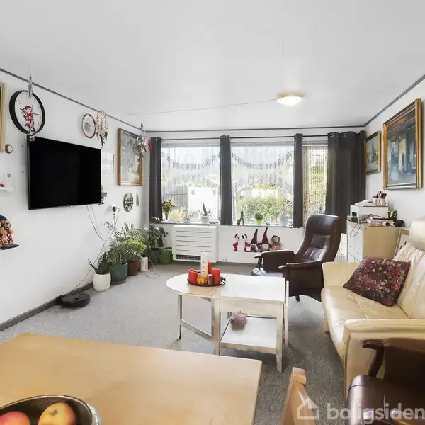Stue med en lys sofa og lænestol omkring et sofabord. Tv hænger på væggen over hylder med dekorationer. Store vinduer giver udsigt til haven. Bord med frugtskål i forgrunden.