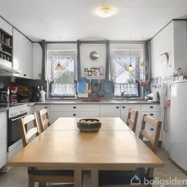 Et køkken med en træspisebord i midten er omgivet af seks stole. På bordet står en sort skål. Køkkenet har hvide skabe, et komfur, en rød mikrobølgeovn og vinduer med gardiner.