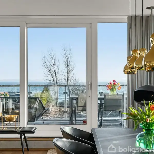 Bord med vase og blomster står ved siden af gyldne lamper over spiseområdet, mens store vinduer viser udsigt til terrasse og havet.