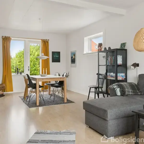 En stue med gråt hjørnesofa står ved siden af et sofabord. Ved spisebordet er der fire stole. Et vindue og åben dør viser en grøn have udenfor. En pejs brænder i hjørnet.