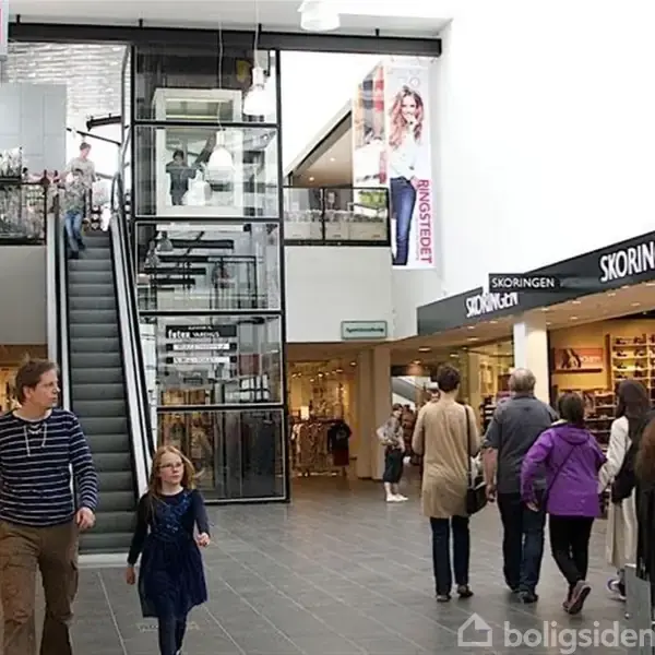 En rulletrappe kører op i et indkøbscenter, omgivet af mennesker. Der er butikker som H&M og Skoringer. Store glasfacader og naturligt lys præger området.