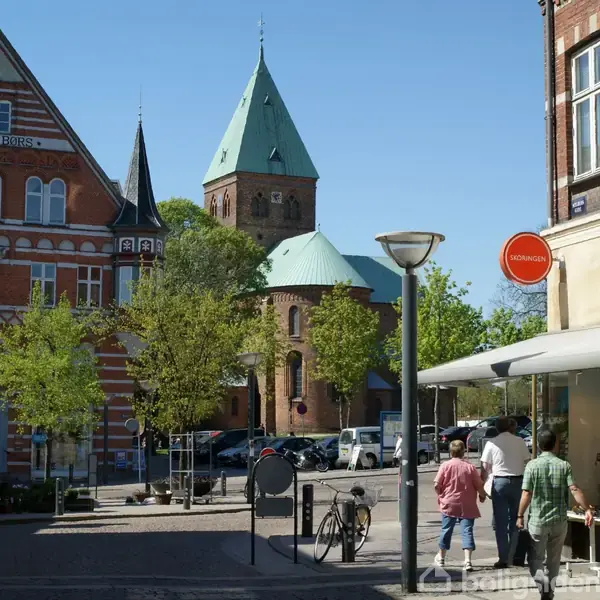 En kirke med grønne tage står omkranset af træer i en bymidte. To personer går forbi en butik under et skilt med teksten "DOMINO". Der er flere cykler parkeret.