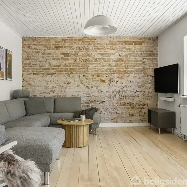 Grå sofa står mod en murstensvæg. Et lille rundt bord står foran sofaen. Fjernsyn hænger på væggen ved siden af et vindue med rullegardin. Tre plakater dekorerer den hvide væg.
