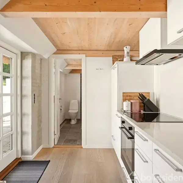Køkken med hvide skabe og komfur, knive i blok på bordplade. Glasdøre åbner til terrasse. Træloft og trægulv. Dør til badeværelse i baggrunden.