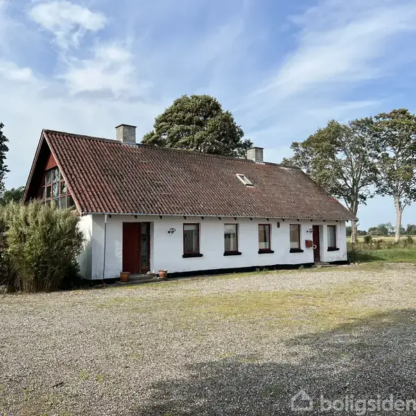 Hvidmalet hus med rødt tag står stille på en grusplads omgivet af buske og træer under en blå himmel.