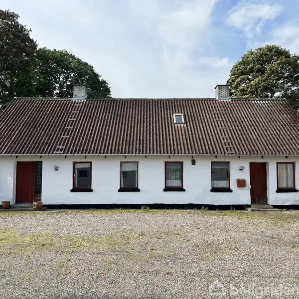 Et hvidt hus med rødt tegltag og mørke vinduer står stille på en grusbelagt gårdsplads omgivet af træer. To døre og fem vinduer er synlige frontalt.