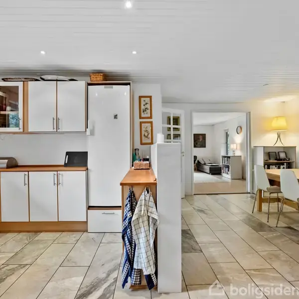 Køkken med hvide skabe og trækanter har en integreret hvid fryser. Viskerhåndklæder hænger på væggen. I baggrunden en spiseplads med bord, stole og lampe ved siden af kunstværk.