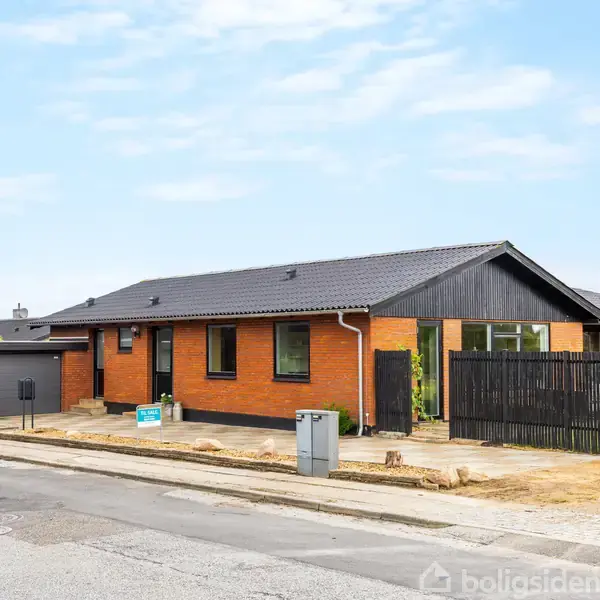 En rød murstensbungalow med sort tag står ved en asfaltvej. Der er en tilhørende garage og sort hegn i forhaven. Himlen er overskyet.