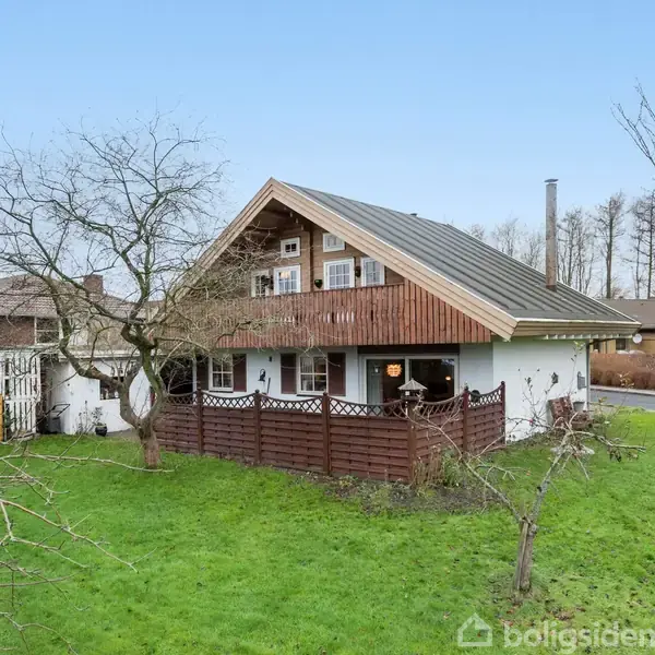 Et hus i træ- og murstensmateriale med en stor, stejl taghældning står blandt nøgne træer på en grøn plæne. En vej ses i baggrunden.