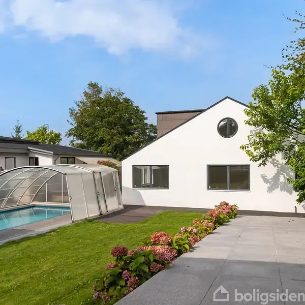 Hus med hvid facade og runde vinduer, står ved siden af en overdækket swimmingpool. Haven er grøn med blomsterbede, omgivet af andre moderne huse og træer under en blå himmel.