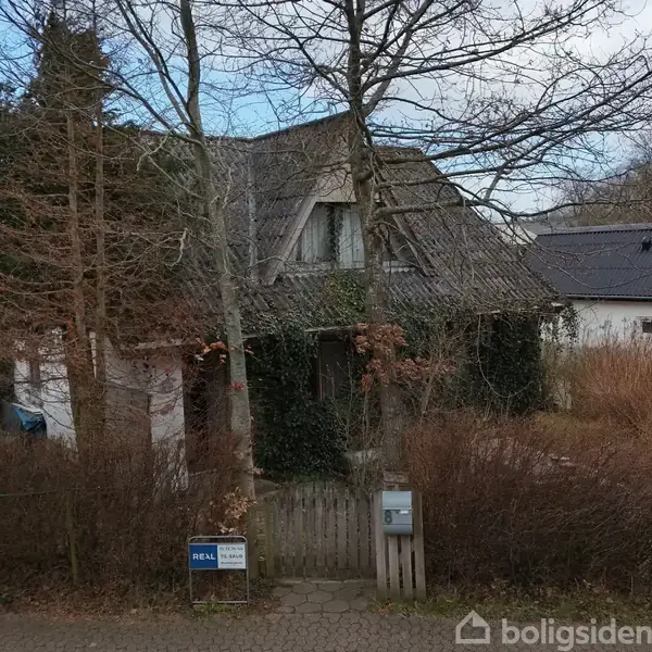Et gammelt hus med et stejl tag er dækket af klatreplanter. En tilgroet have omkranser det. Ved indgangsporten ses et skilt med teksten: "TIL SALG" og "EDC".