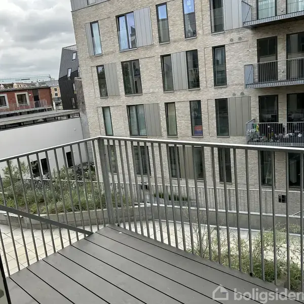 En balkon med metalrækværk er roligt placeret, hvor den giver udsigt til en moderne, grå murstensbygning med store vinduer og altaner. Grønne planter pryder området omkring bygningen.