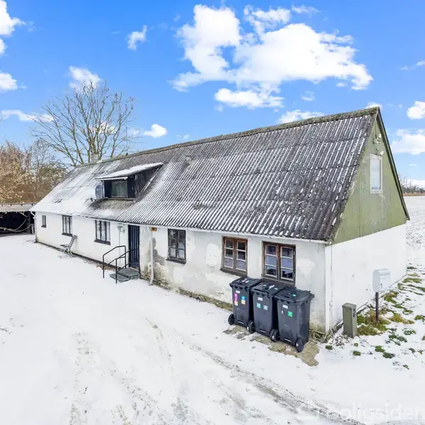 Et hvidt landhus med sort tag render gennem en snedækket landskab. Foran bygningen står tre affaldscontainere. Træer og marker omkranser huset under en blå himmel med skyer.