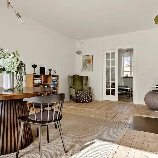 Rundt spisebord med fire træstole står i et lyst rum. En kamin er tændt ved siden af en grøn stol og et reolsystem. Store vinduer slipper lys ind i rummet.