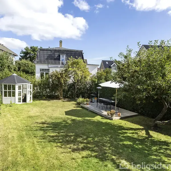En velplejet have med et lyst havehus står stille. Omkring er der frodig beplantning, og en terrasse er dækket af en parasol. Baggrunden viser huse under en blå himmel.