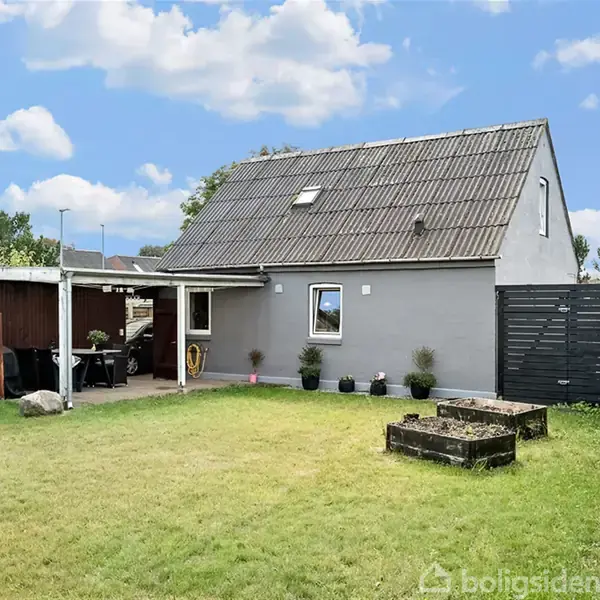 Et gråt hus står stille i en grøn have med blomsterkrukker foran. Til venstre er en overdækket terrasse med møbler. Skyer og blå himmel i baggrunden.