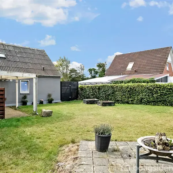 Et gråt hus står i en have med græsplæne. En bålplads er placeret på fliserne i forgrunden, omgivet af et hegn og flere huse i baggrunden.