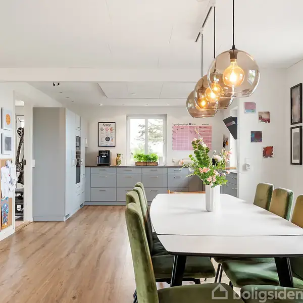 Spisebord med grønne stole står under moderne lamper. En pejs brænder i hjørnet. Køkkenet har grå skabe og planter i vinduet. Trægulv gennem hele rummet.