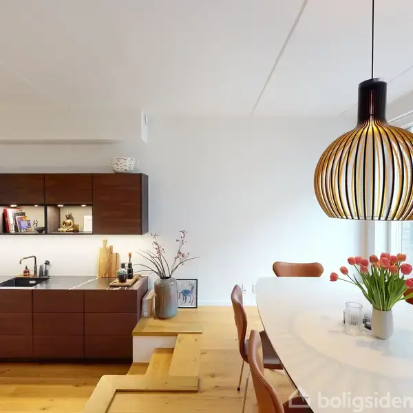 Et moderne spisebord med en hvid bordplade er omgivet af seks brune stole. En dekorativ lampe hænger over bordet, der står i et lyst rum med træstrapper i baggrunden.