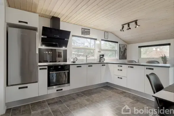 Et moderne køkken med hvide skabe og stålapparater står ryddeligt. På væggen hænger teksten: "Et køkken uden rod er som..." Rummet har lyst træloft og flisegulv.