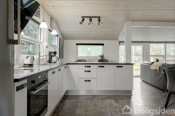 Køkken med hvide skabe står i et lyst rum. Kaffemaskine og elkedel er på bordet, mens store vinduer giver udsigt til en have.